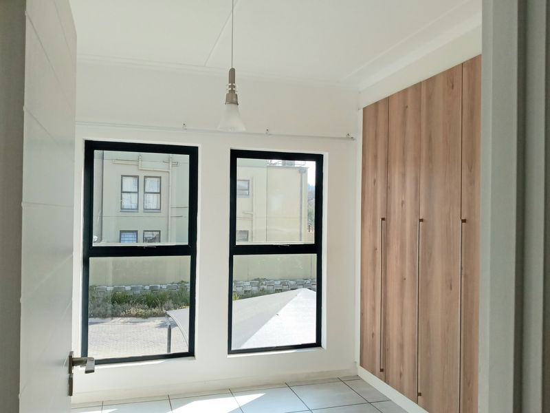 A bright, modern bedroom featuring neutral tiled flooring, sleek built-in wooden cupboards, and large aluminium-framed windows providing ample natural light.