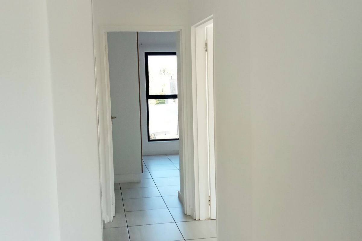 A clean, bright hallway featuring neutral tiled flooring and white walls, leading towards an adjacent room with natural light.