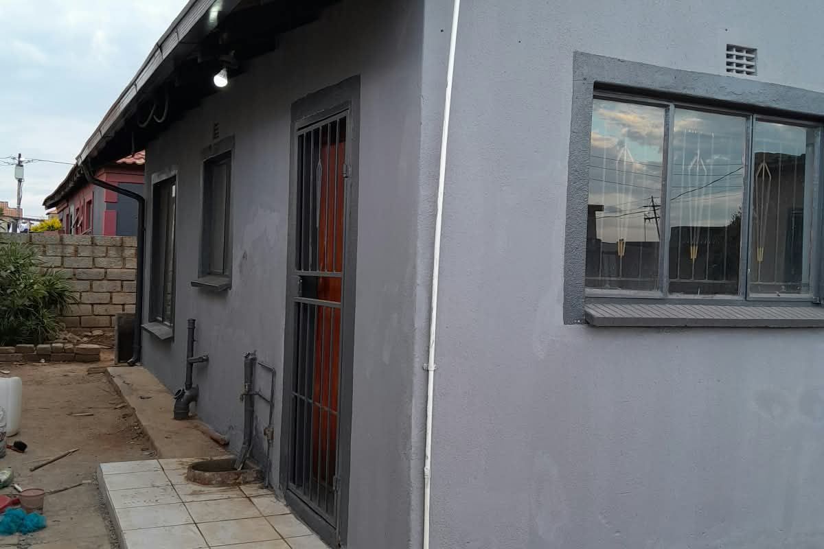 Rear exterior view of the house featuring a tiled walkway, security gate, and grey painted facade with metal window frames.