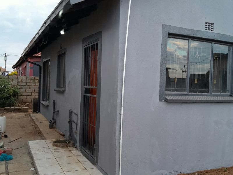 Rear exterior view of the house featuring a tiled walkway, security gate, and grey painted facade with metal window frames.