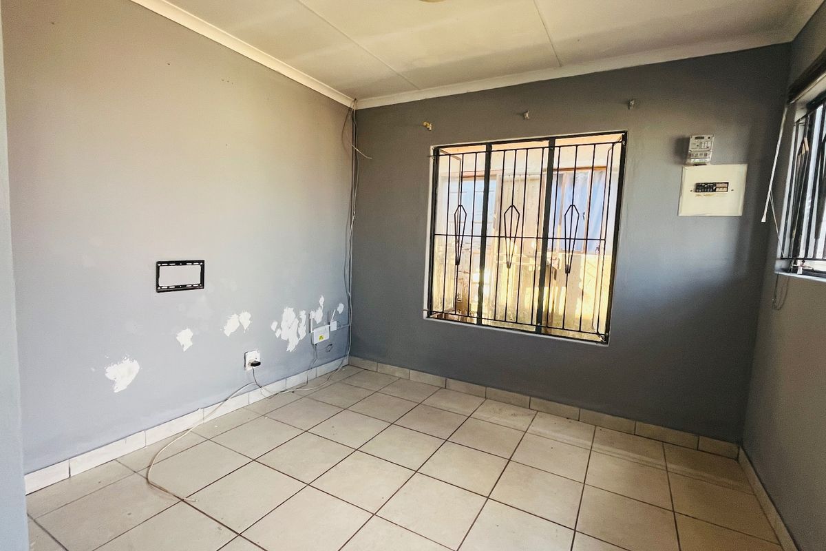 An empty living room featuring tiled flooring, security bars on windows, and an electrical distribution board, requiring cosmetic wall repairs.