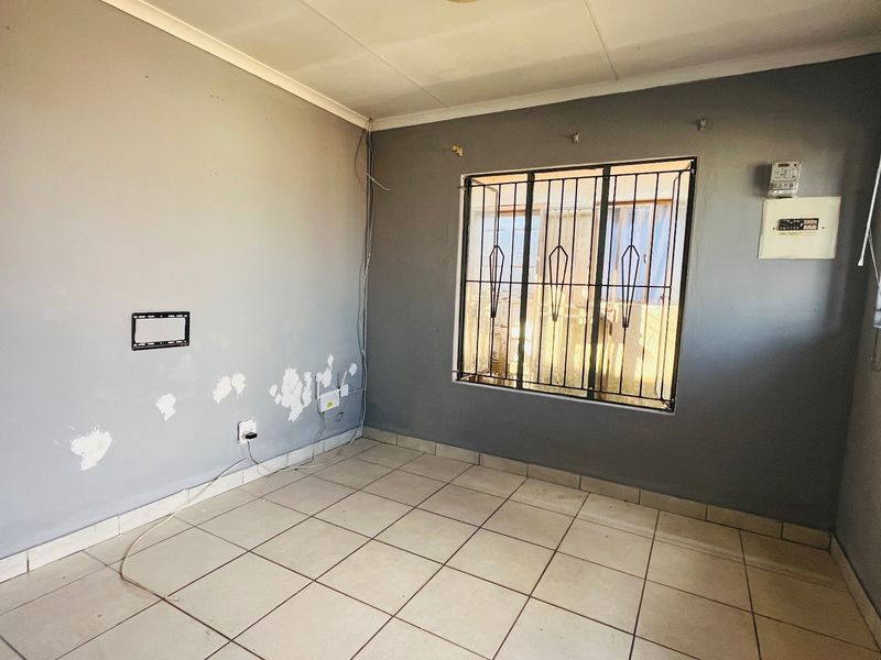 An empty living room featuring tiled flooring, security bars on windows, and an electrical distribution board, requiring cosmetic wall repairs.