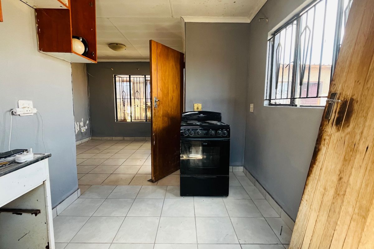 A basic kitchen area featuring tiled floors, a freestanding stove, and security bars on the windows, currently in need of cosmetic updates and repairs.