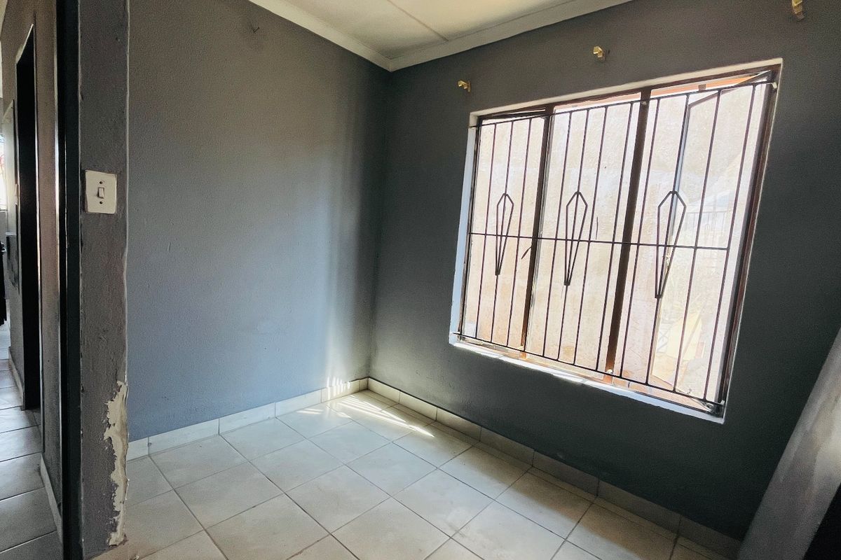 An empty bedroom featuring tiled flooring, grey painted walls, and security bars on the window. The space requires minor cosmetic repairs to the wall plaster.