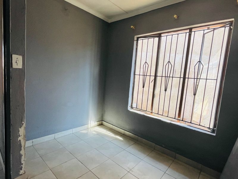 An empty bedroom featuring tiled flooring, grey painted walls, and security bars on the window. The space requires minor cosmetic repairs to the wall plaster.