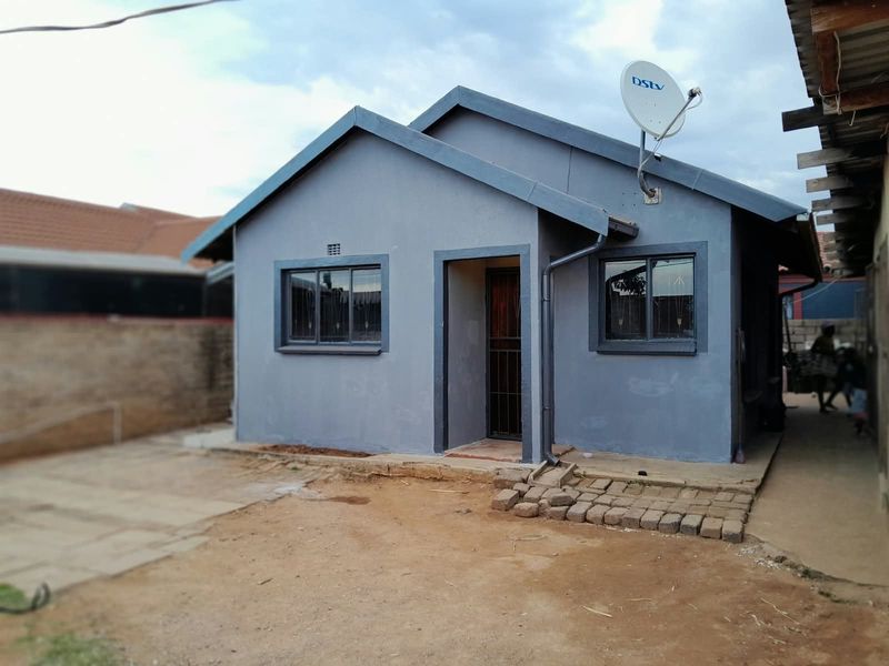 A single-story residential house featuring a grey painted facade, aluminium window frames, and a paved entrance area.