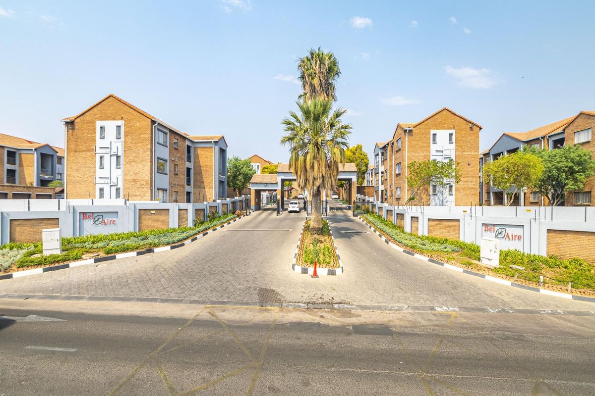 The entrance to Bel Aire apartment complex featuring a paved driveway, secure gated access, brick facade buildings, and well-maintained perimeter walls with electric fencing.