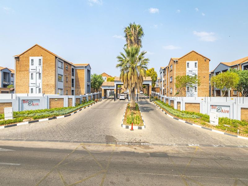 The entrance to Bel Aire apartment complex featuring a paved driveway, secure gated access, brick facade buildings, and well-maintained perimeter walls with electric fencing.