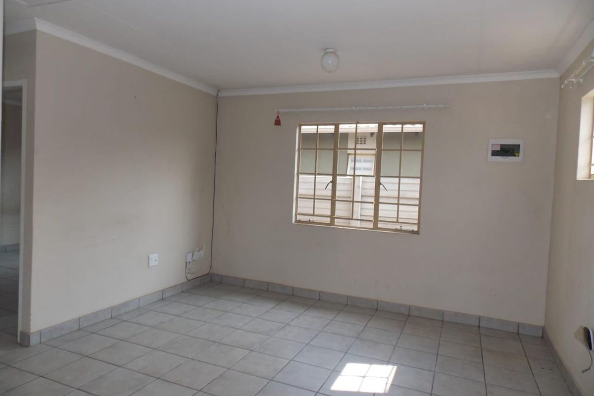 An empty living room featuring neutral tiled flooring, white walls with cove cornicing, and a window fitted with security burglar bars.