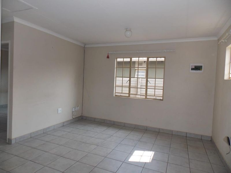 An empty living room featuring neutral tiled flooring, white walls with cove cornicing, and a window fitted with security burglar bars.