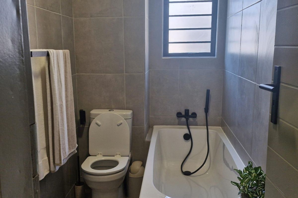 A modern bathroom featuring grey wall tiles, a bathtub with a black shower mixer, and a louvered window for ventilation.