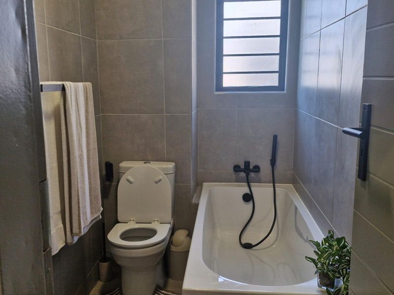A modern bathroom featuring grey wall tiles, a bathtub with a black shower mixer, and a louvered window for ventilation.