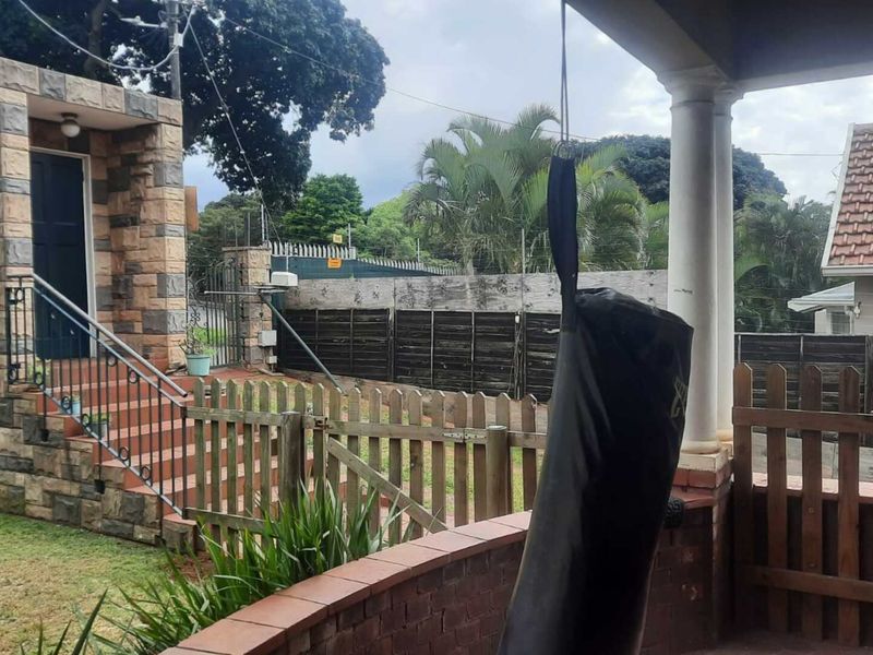 A covered patio area featuring stone cladding, concrete columns, and a wrought iron railing, overlooking a fenced garden and exterior entrance.