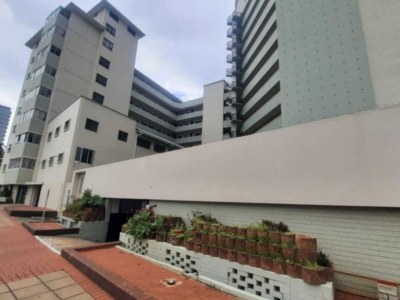 Exterior view of a multi-story apartment building featuring a perimeter wall, paved walkway, and access to underground parking.