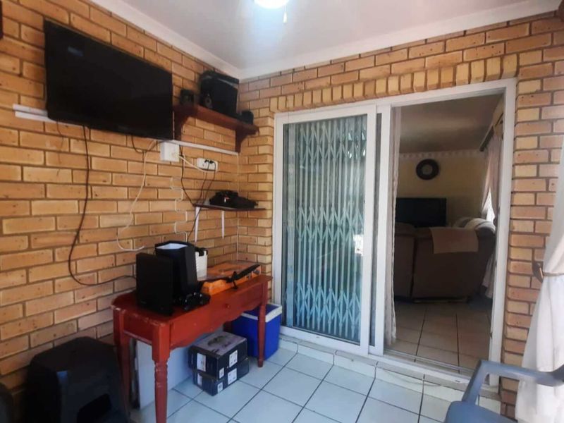 A covered patio area featuring exposed brick walls, tiled flooring, and a sliding glass door with a security gate providing access to the interior.