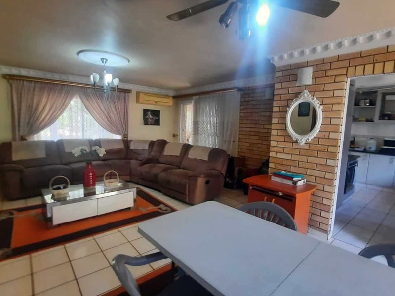 Open plan living and dining area featuring exposed brickwork, tiled flooring, and a ceiling fan, with an adjacent kitchen visible through the doorway.
