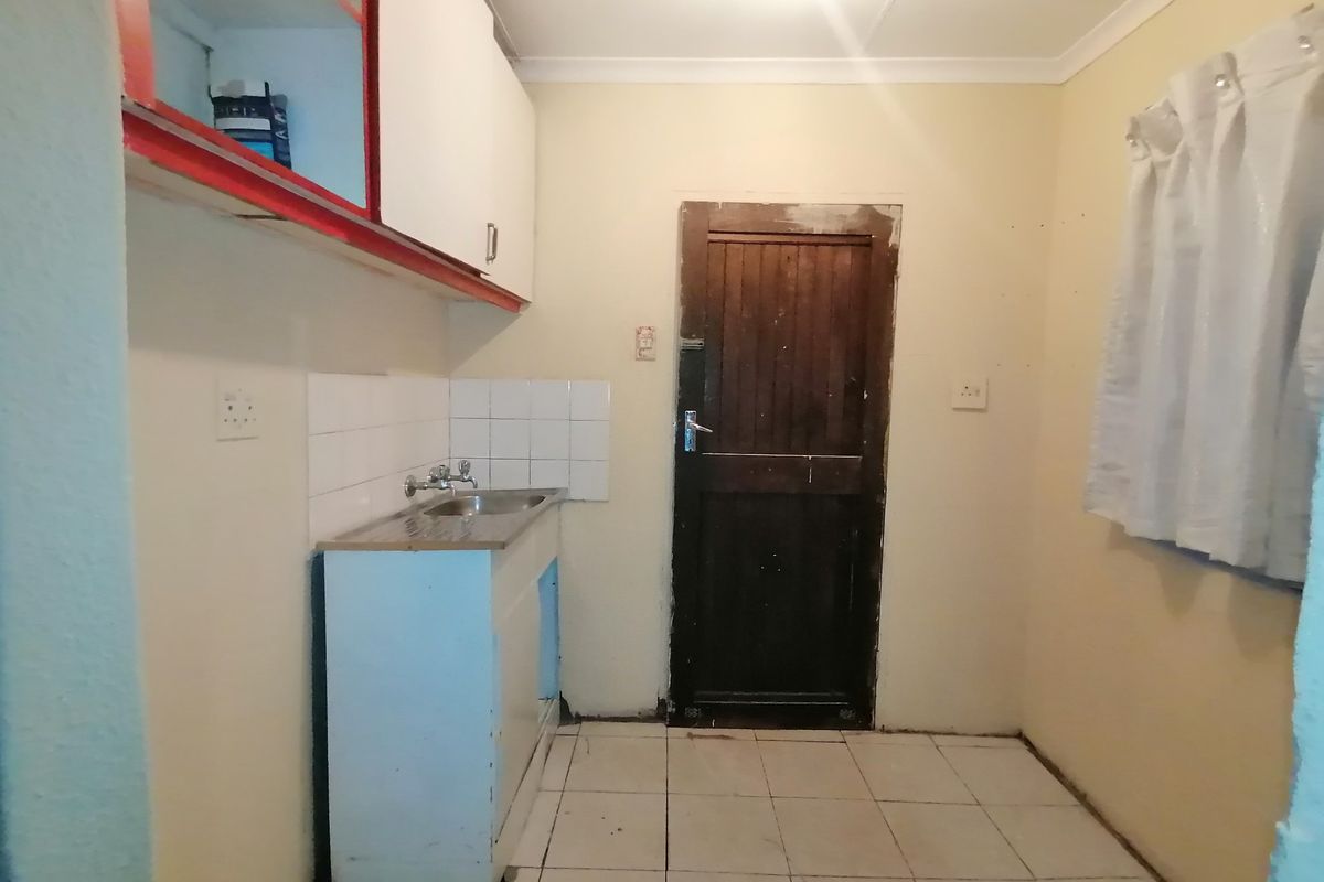 A small, dated kitchen area featuring basic cabinetry, a stainless steel sink, and tiled flooring, requiring significant cosmetic updates and repairs.