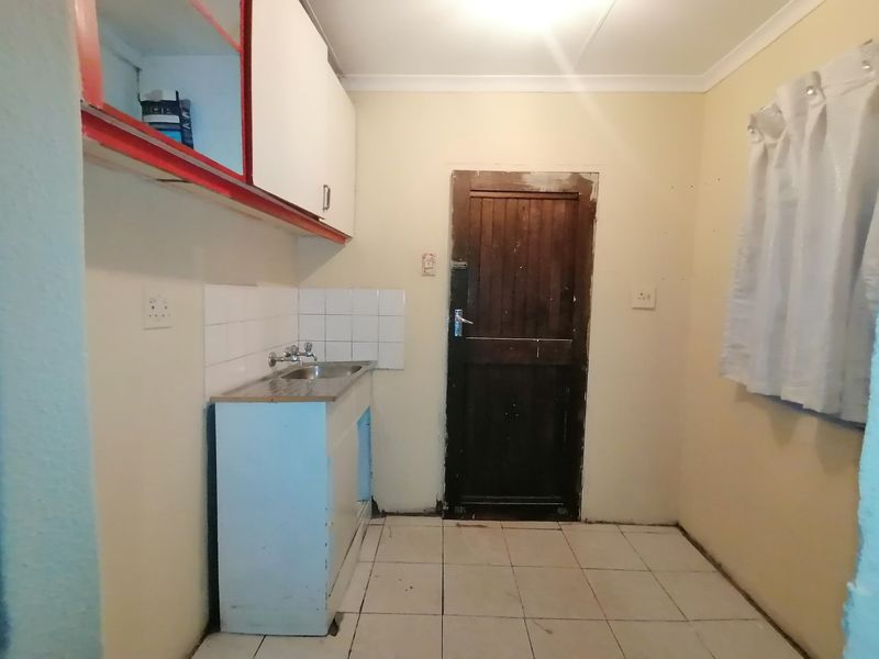 A small, dated kitchen area featuring basic cabinetry, a stainless steel sink, and tiled flooring, requiring significant cosmetic updates and repairs.