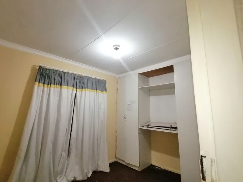 A compact bedroom featuring a built-in wardrobe with open shelving and standard ceiling lighting, requiring cosmetic updates to improve its overall condition.
