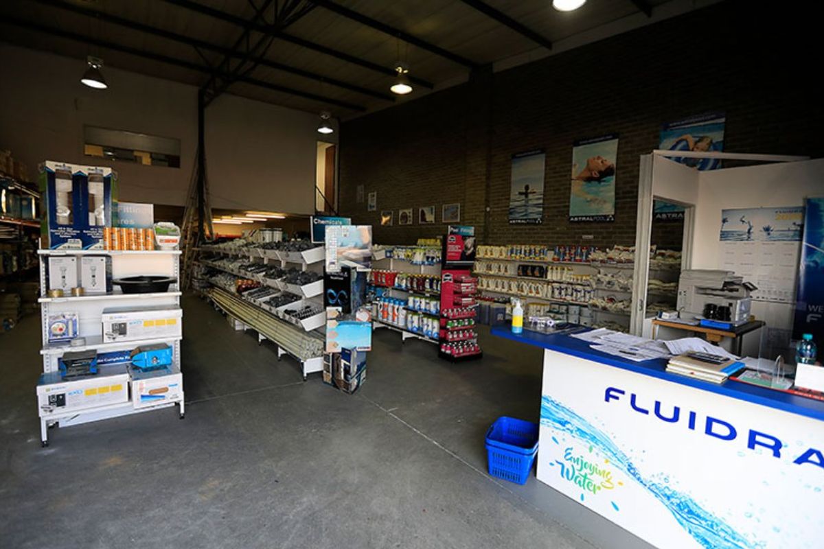 Commercial retail space featuring exposed brick walls, industrial steel roof trusses, concrete flooring, and a mezzanine level, currently utilized for pool supply inventory.