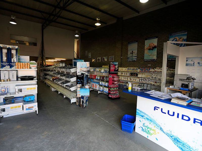 Commercial retail space featuring exposed brick walls, industrial steel roof trusses, concrete flooring, and a mezzanine level, currently utilized for pool supply inventory.