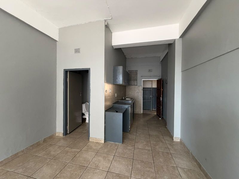 Open plan living area featuring tiled flooring, a compact kitchenette with built-in cabinetry, and access to an adjacent bathroom and secondary entrance.