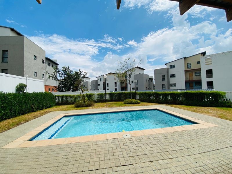 A well-maintained communal swimming pool area featuring paved surrounds and manicured hedging within a modern apartment complex.