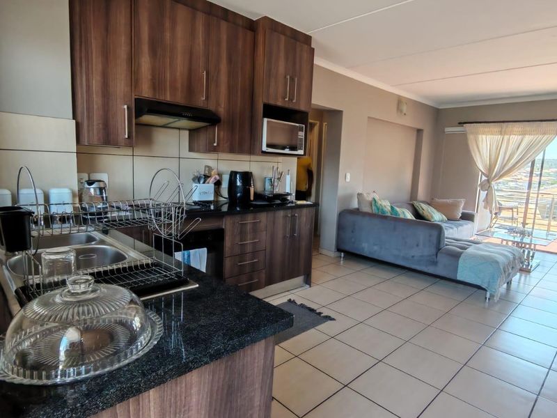 Open-plan kitchen and living area featuring dark wood cabinetry, granite countertops, and tiled flooring, leading to a bright living space with balcony access.