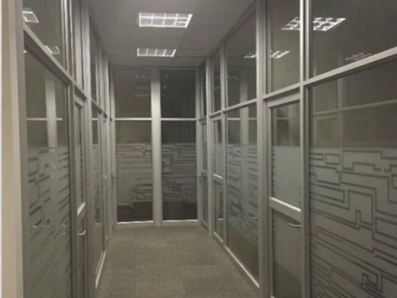 A commercial office hallway featuring aluminum-framed glass partitions, recessed lighting, and carpeted flooring, providing a clean and professional workspace layout.