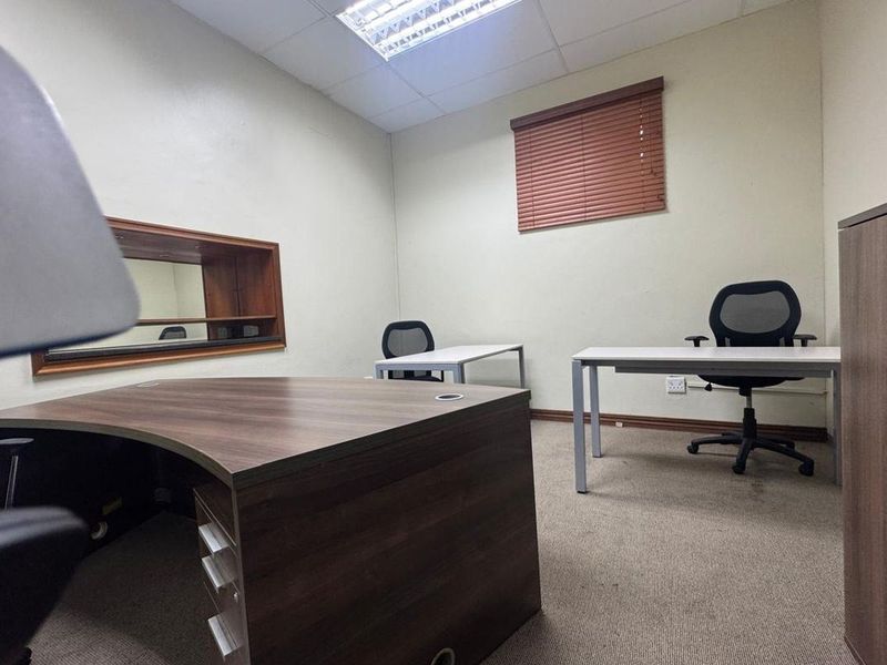 A functional commercial office space featuring carpeted flooring, a suspended ceiling with recessed lighting, and an internal window with wooden blinds.