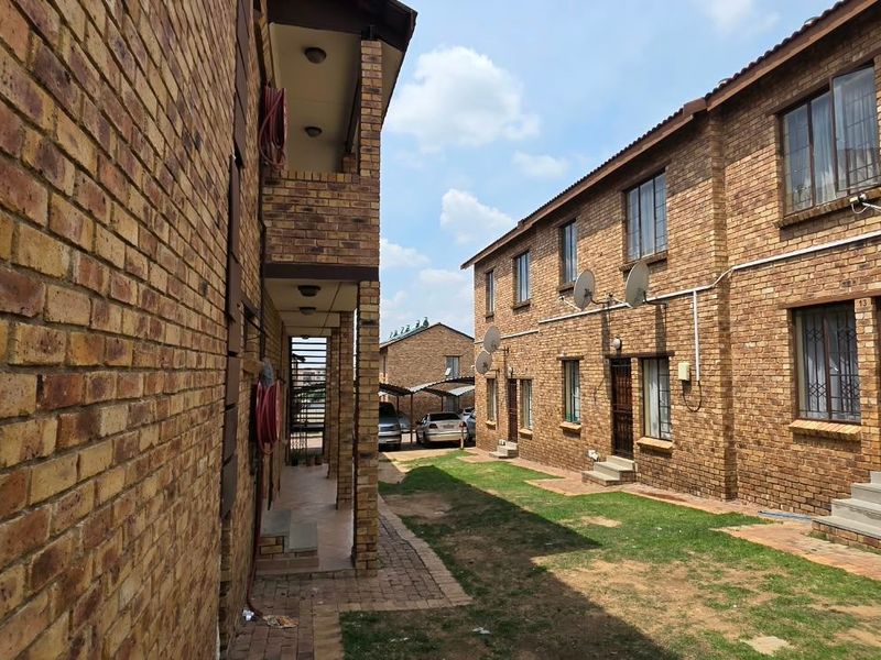 Exterior view of a face brick apartment complex featuring communal garden space, paved walkways, and security fixtures on windows and doors.
