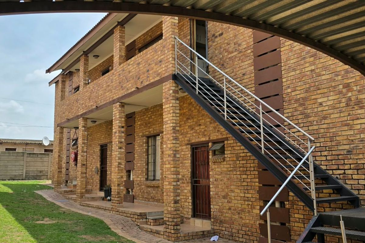 Exterior view of a two-story face brick apartment building featuring an external metal staircase with stainless steel handrails and a paved garden area.