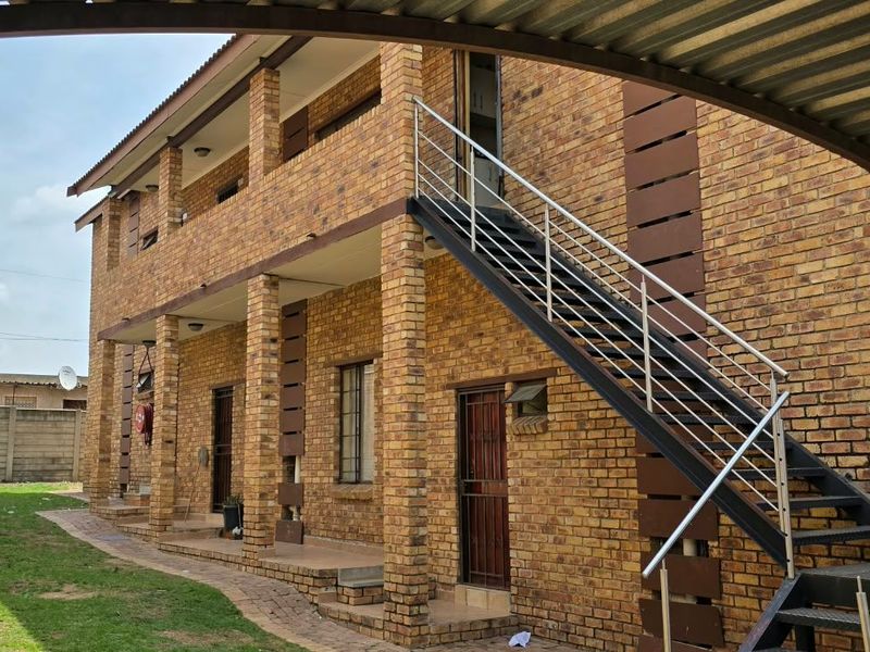 Exterior view of a two-story face brick apartment building featuring an external metal staircase with stainless steel handrails and a paved garden area.