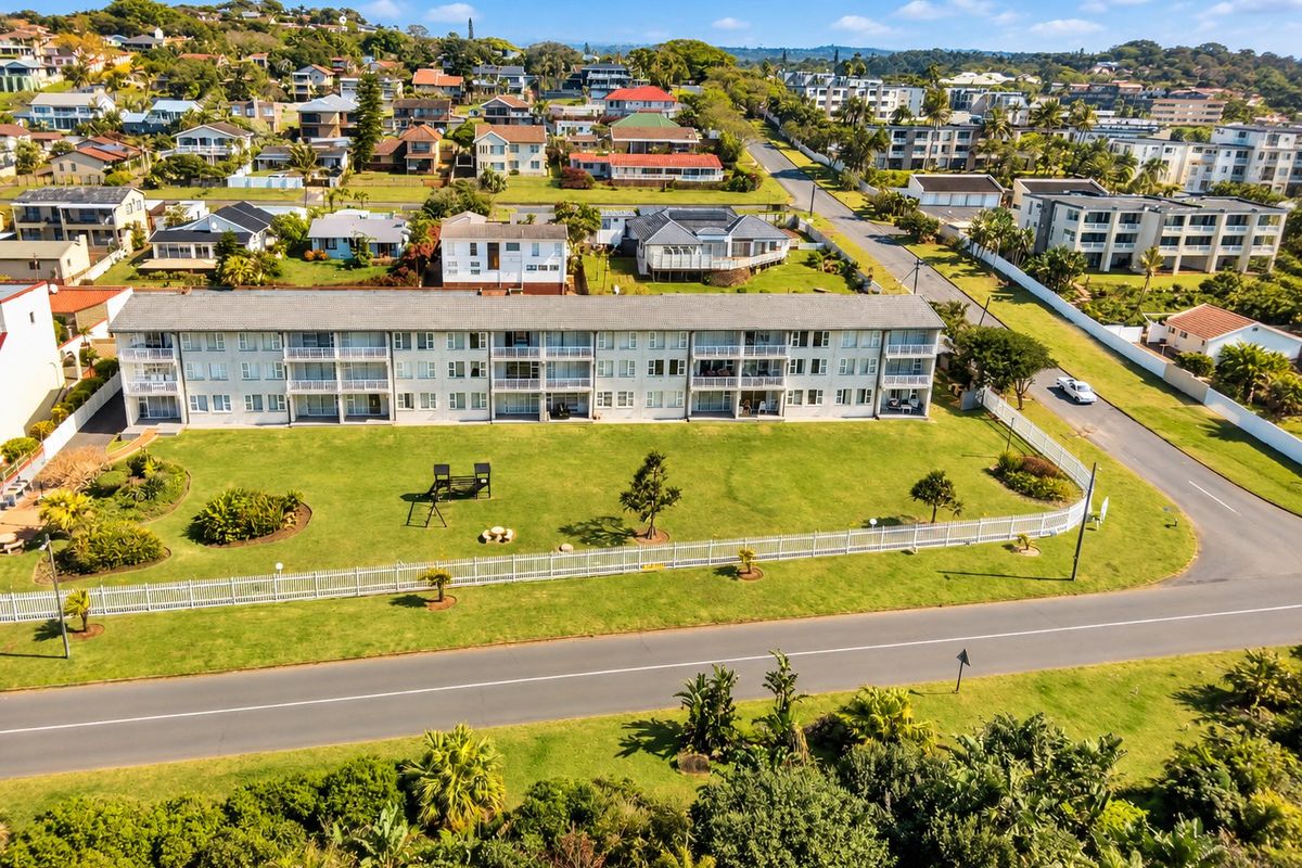 Aerial view of a multi-story apartment complex featuring a large communal lawn, white picket fencing, and private balconies for each unit.