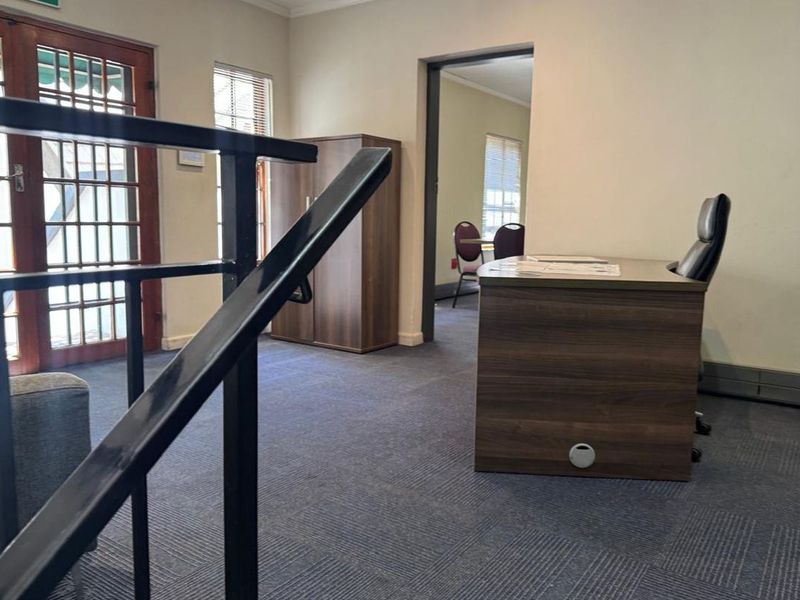 A functional office space featuring carpeted flooring, a timber-framed glass door leading to an outdoor area, and an internal doorway connecting to an adjacent room.