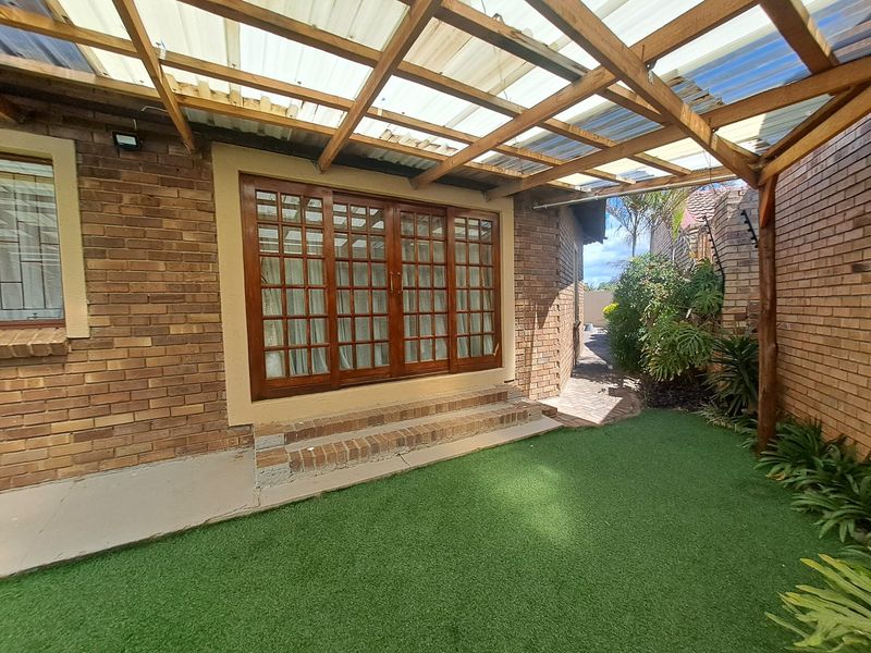 A covered patio area featuring a timber pergola with polycarbonate roofing, facebrick walls, and access via timber sliding doors to an artificial lawn garden.