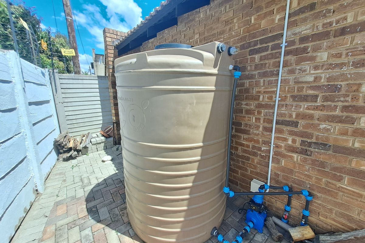 Exterior rear area featuring a large water storage tank with an integrated pump system installed against a brick wall on paved ground.
