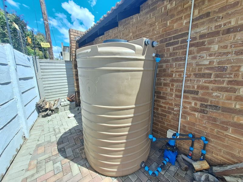 Exterior rear area featuring a large water storage tank with an integrated pump system installed against a brick wall on paved ground.