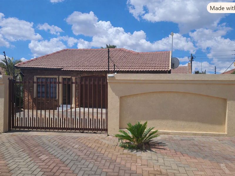 Front view of a single-story brick house featuring a tiled roof, secure perimeter wall, and a paved driveway with a metal gate.