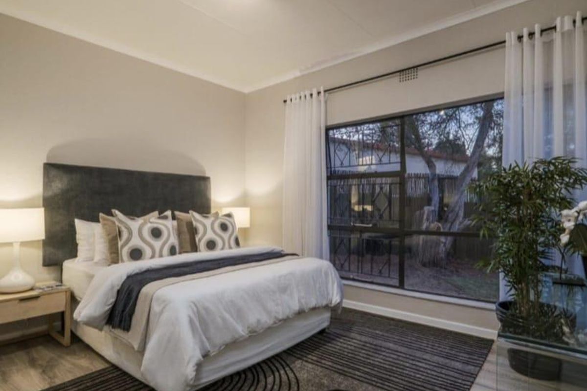 A well-maintained bedroom featuring laminate flooring, neutral wall tones, and a large window with security bars overlooking the garden.