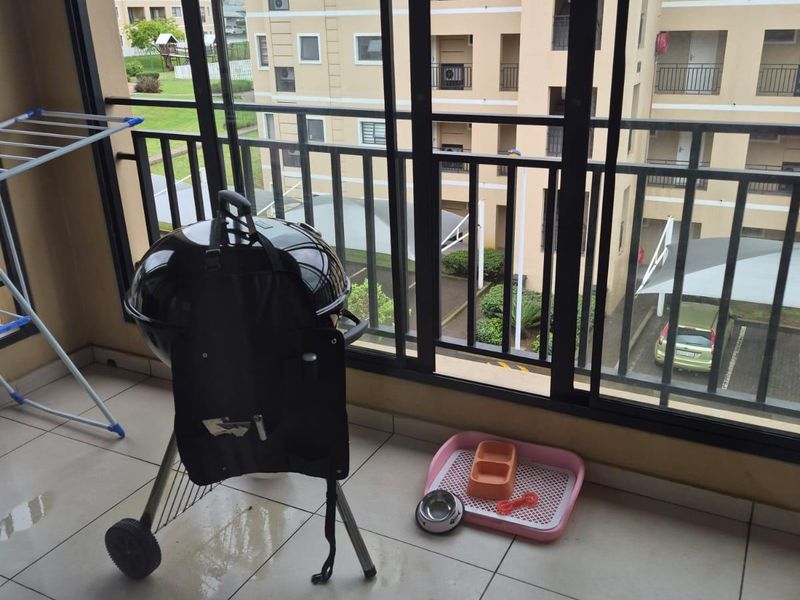 A covered balcony featuring tiled flooring, black aluminium framed windows, and a metal balustrade overlooking the apartment complex grounds.