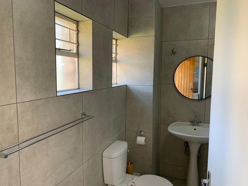 A clean, modern bathroom featuring neutral wall tiling, a pedestal basin, and a standard toilet with natural light from casement windows.