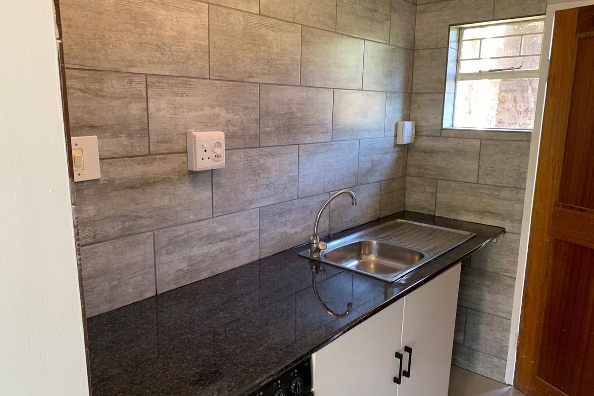A compact kitchen area featuring a granite countertop, stainless steel sink, built-in oven, and modern grey tiled backsplash.