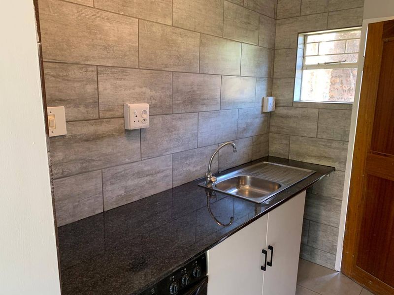 A compact kitchen area featuring a granite countertop, stainless steel sink, built-in oven, and modern grey tiled backsplash.