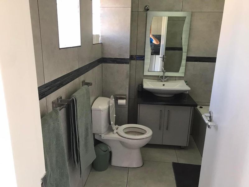 A functional bathroom featuring a modern vanity with basin, wall-mounted mirror, toilet, and neutral grey wall and floor tiling.