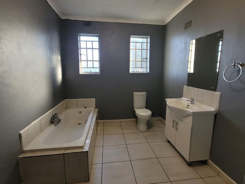 A spacious bathroom featuring a built-in bathtub, vanity cabinet with basin, and toilet, finished with tiled flooring and dark grey walls.