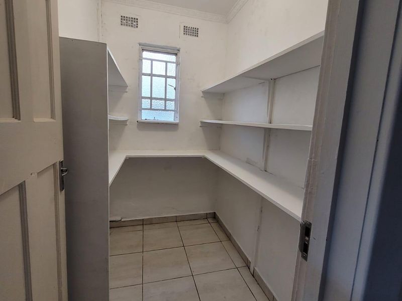 A functional scullery or pantry area featuring built-in shelving, tiled flooring, and a small window with security bars.