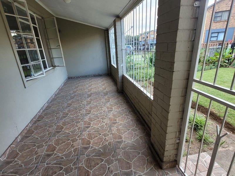 A covered patio area featuring tiled flooring, brick pillars, and security bars, offering a view of the garden and surrounding townhouse complex.