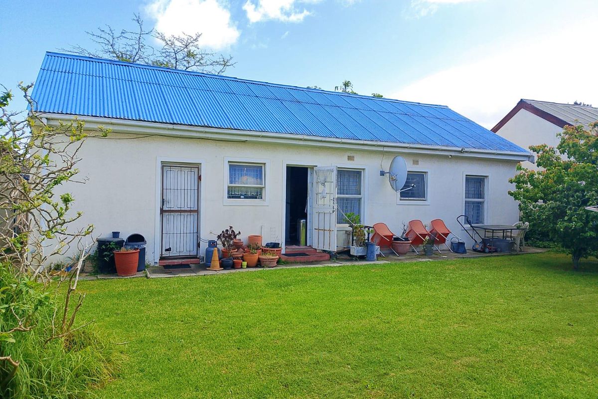 Rear view of a single-story house featuring a blue corrugated metal roof, white exterior walls, and a paved patio area leading to a garden.
