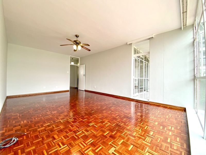 Spacious living area featuring polished parquet flooring, a ceiling fan, and large windows that provide ample natural light.
